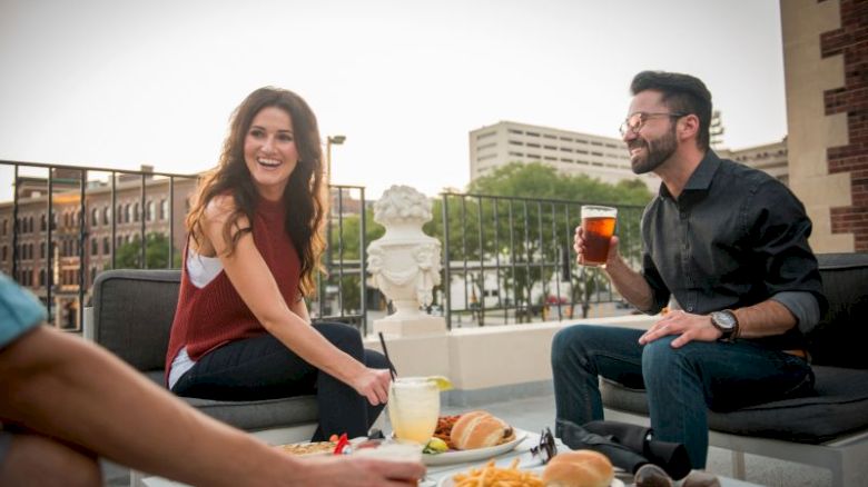 A group of people enjoying drinks and food on a rooftop patio, with city buildings in the background.