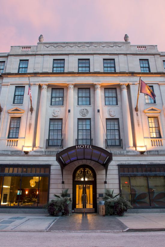 The image shows a grand building with flags, large windows, and an entrance canopy, likely a hotel or historical structure at sunset.