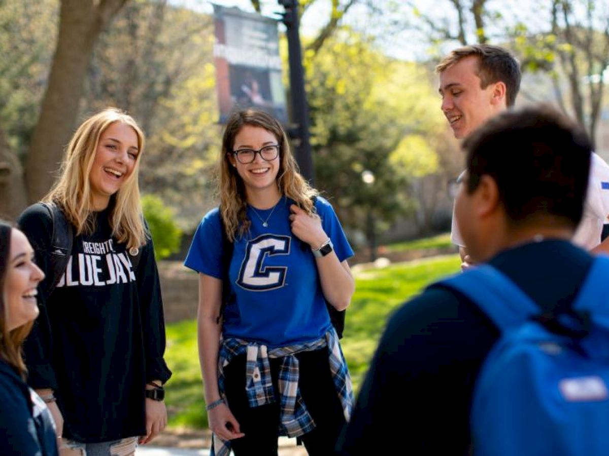 A group of students is outside, smiling and talking on a sunny day, with trees and grass in the background, wearing casual clothing.