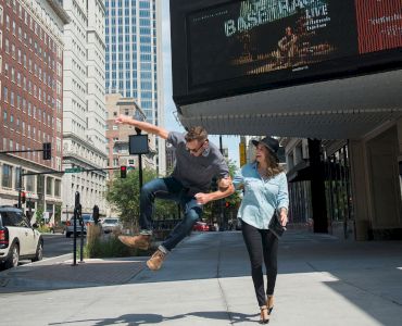 A man jumps playfully next to a woman on a city street with tall buildings and a theater marquee in the background.