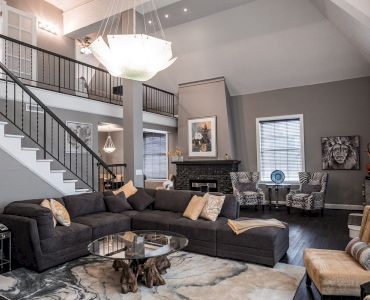 The image shows a modern living room with a gray sectional sofa, art pieces, and a chandelier, beneath a staircase leading to a loft area.