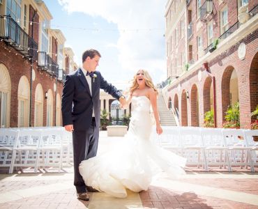 A couple in wedding attire is joyfully celebrating in a picturesque courtyard with empty white chairs and brick buildings in the background.