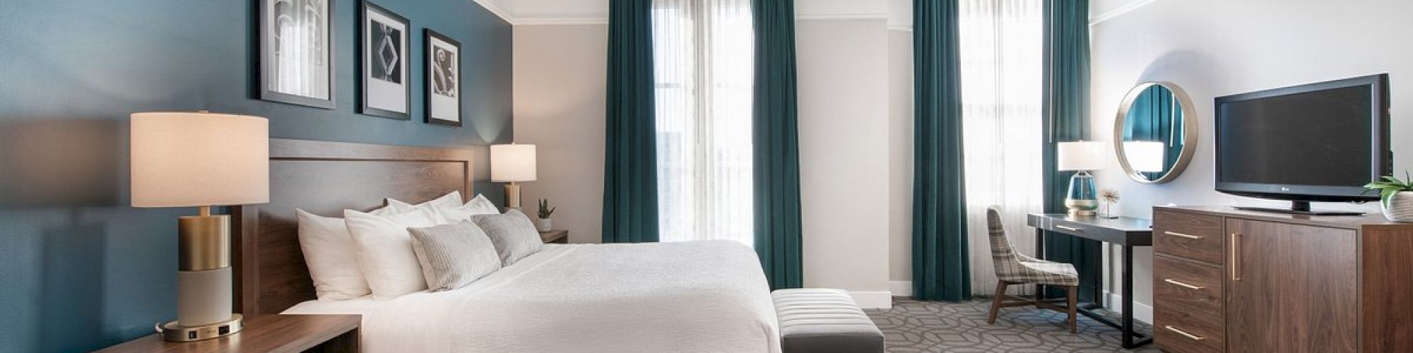 A modern bedroom with a king-sized bed, nightstands, desk, chair, TV, and patterned carpet, all against a blue accent wall.