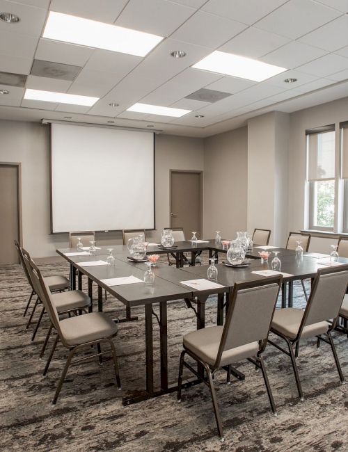 A well-lit conference room with a rectangular table, chairs, a projector screen, and large windows is shown in this image.