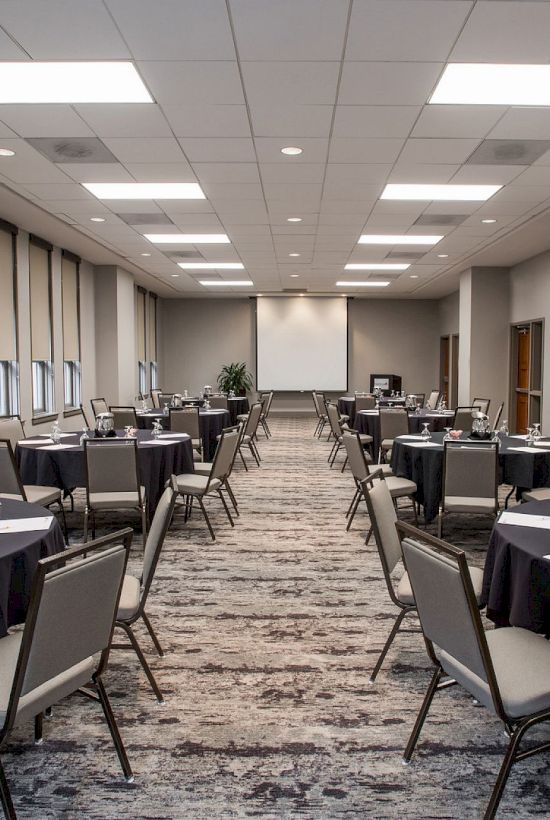 A conference room with round tables, set for a meeting or event; tables have papers, water pitchers, and glasses, leading to a screen.