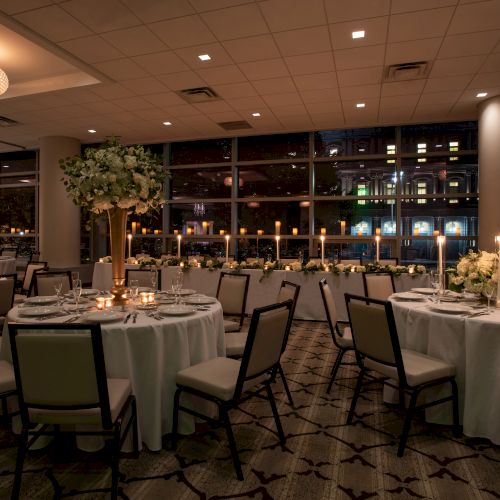 A formal, elegantly set dining room with round tables, white linens, floral centerpieces, and candlelight, likely prepared for an event.