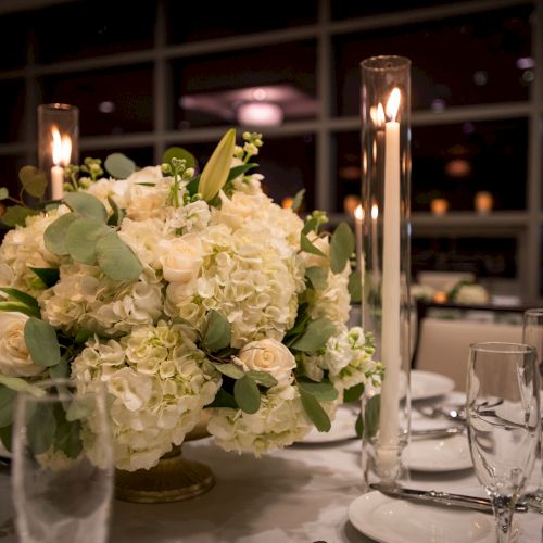 A floral centerpiece with white flowers and greenery on a dining table, surrounded by tall candles and glassware, creating an elegant ambiance.