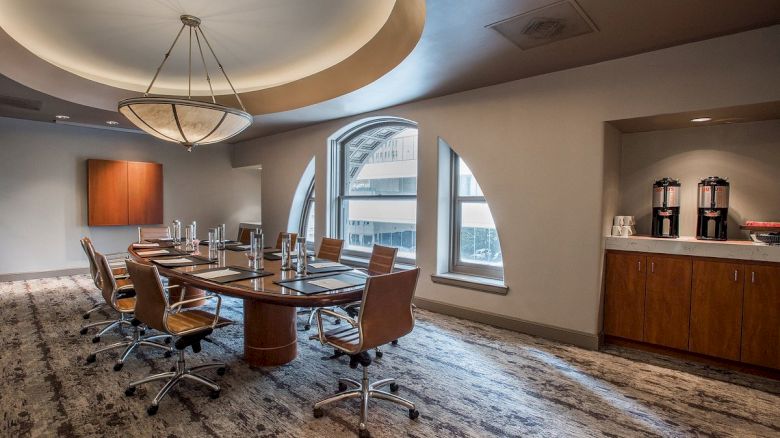 A modern conference room with a long table, office chairs, and a side counter with beverages. It has large windows and a stylish ceiling light.