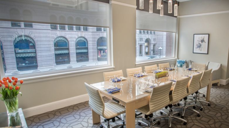 A modern conference room with a long table, chairs, glasses, and napkins, featuring large windows and a city view.