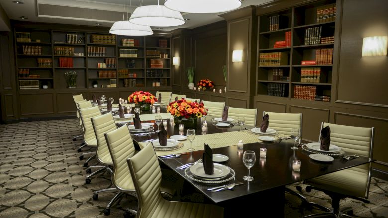 A dining room with a long table, elegant place settings, floral centerpieces, bookshelves, and pendant lighting.