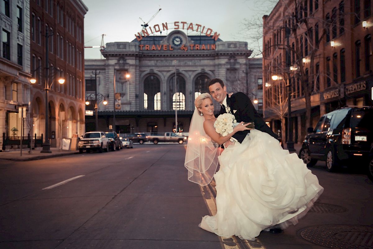 A couple in wedding attire poses happily on a street, with a historic station building in the background.