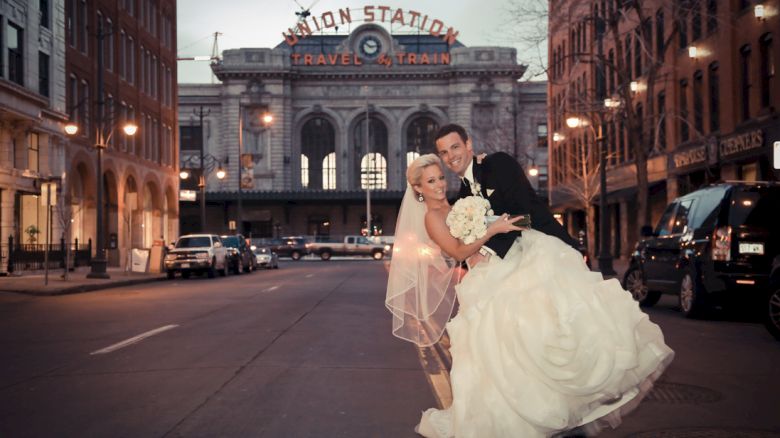 A couple in wedding attire poses happily on a street, with a historic station building in the background.