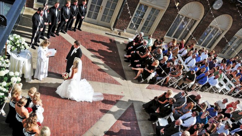 A wedding ceremony is taking place outdoors with a bride, groom, and officiant. Guests are seated, and the wedding party stands nearby.