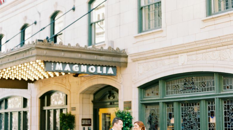 A couple is walking in front of The Magnolia Hotel. The building has flags and decorative elements, suggesting an elegant setting.