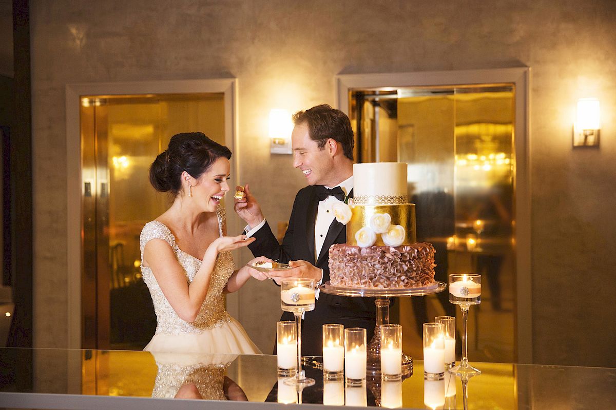 A couple in wedding attire is playfully feeding each other cake in a luxurious setting with candles lit around the table.