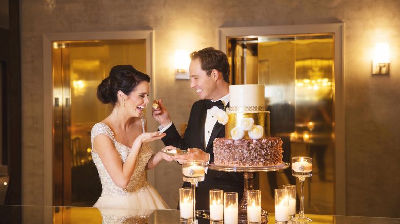 A couple in wedding attire is playfully feeding each other cake in a luxurious setting with candles lit around the table.