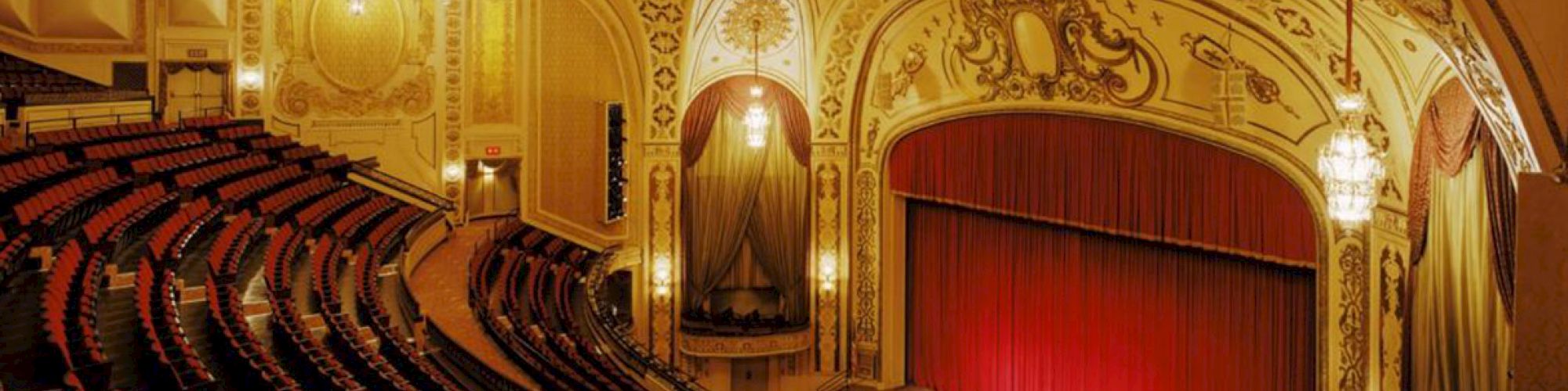 An ornate theater interior with an elegant stage, grand drapes, detailed ceiling, and chandeliers. Rows of empty seats fill the auditorium.