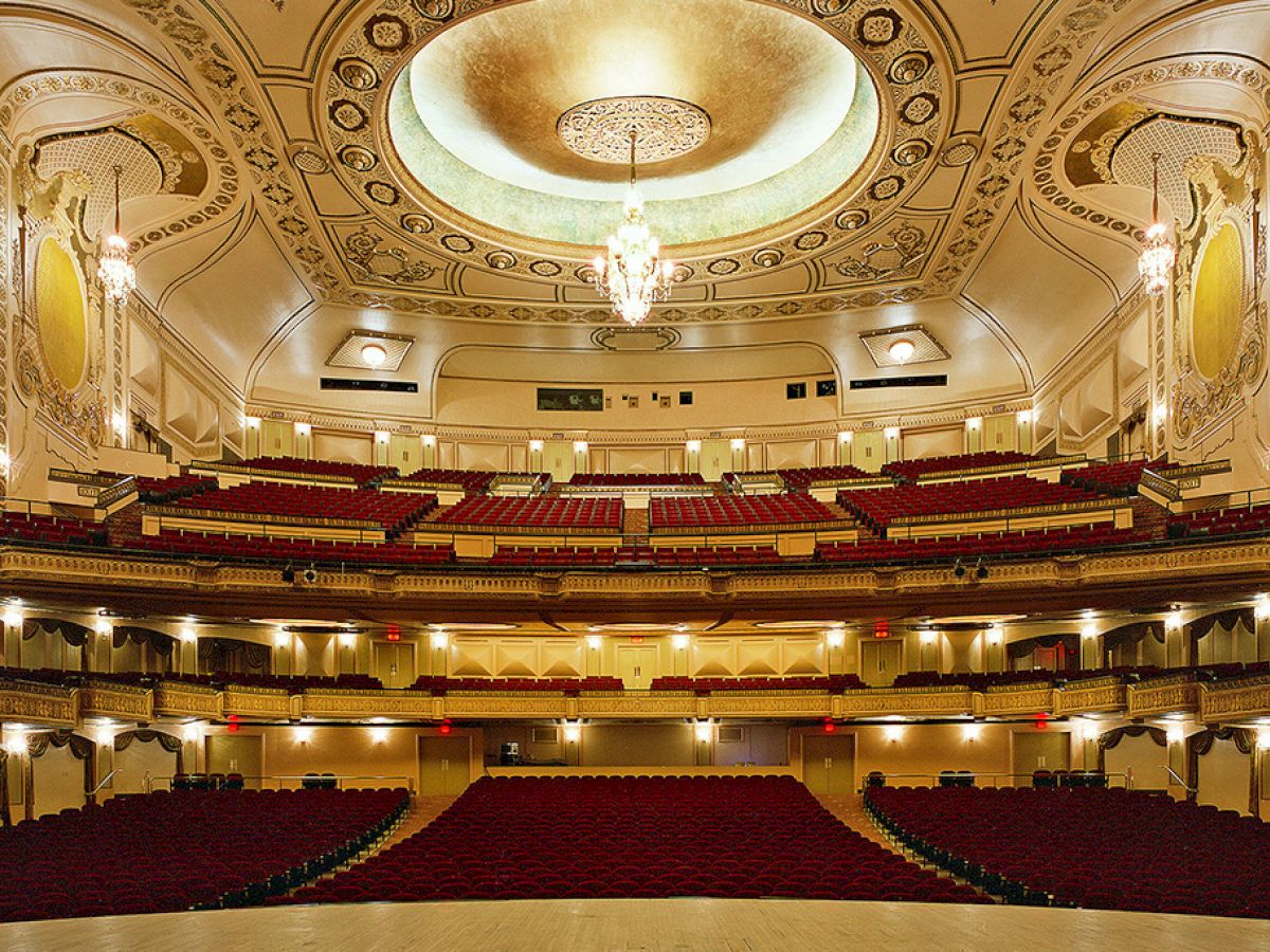 The image shows an ornate, empty theater with multiple seating levels, elegant chandeliers, and decorative details on the ceiling and walls.