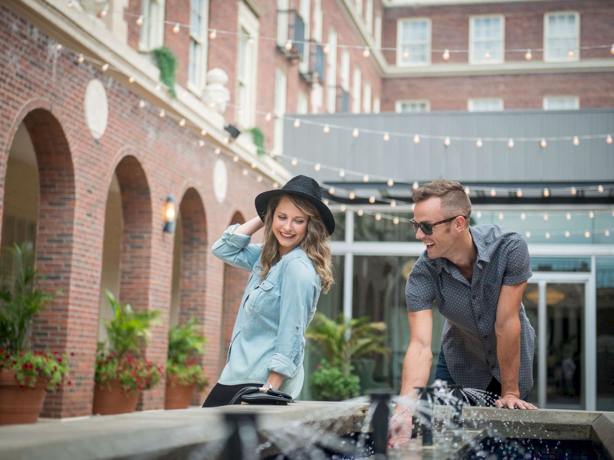 Two people enjoy a playful moment near a water fountain in a courtyard with brick buildings and string lights overhead.