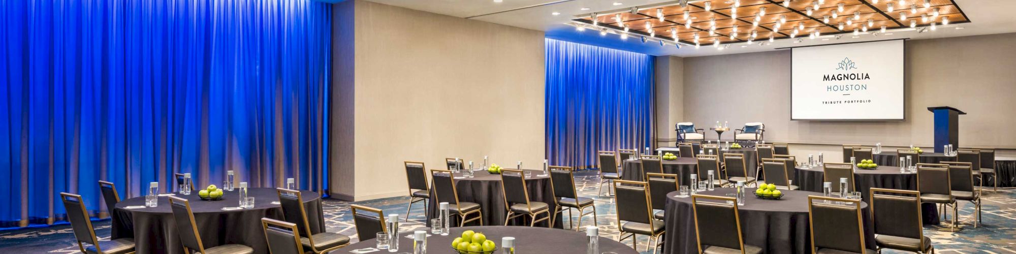 The image shows a modern conference room with round tables, chairs, blue curtains, and a screen displaying 