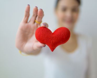 A person is holding a small red heart object in an outstretched hand, with a blurred background.