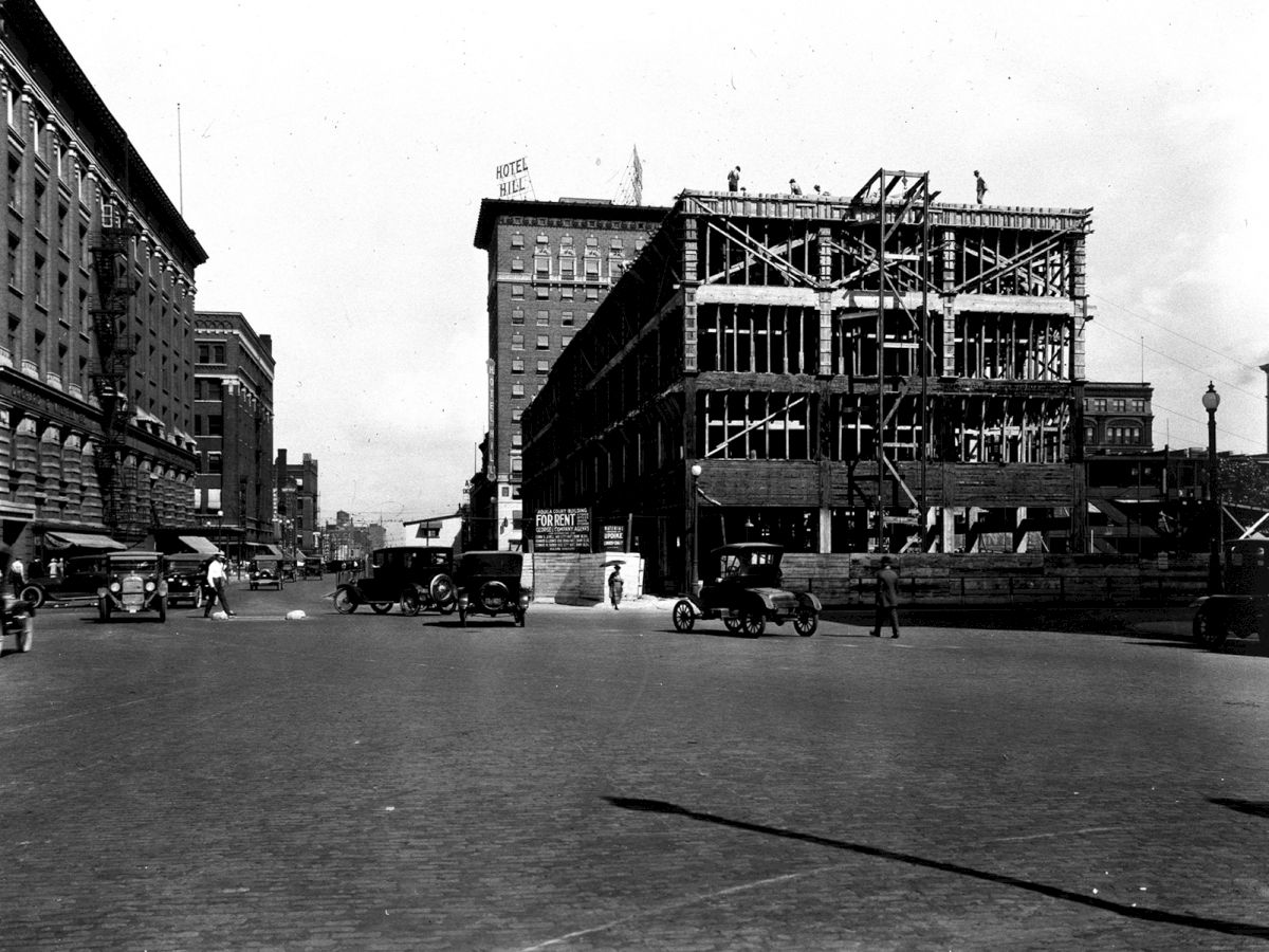 The image shows a historical street scene with early cars, a partially constructed building, and surrounding urban architecture.