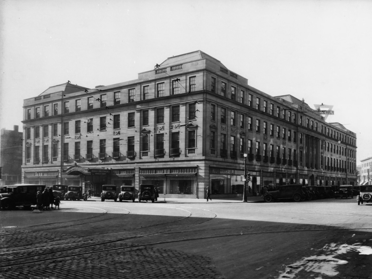 The image shows a large, historic building on a street corner with parked vintage cars in front of it.