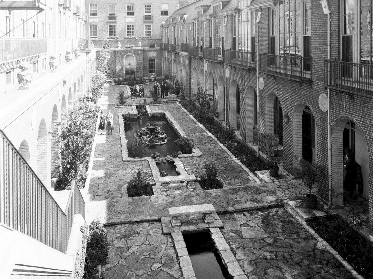 The image shows a historic courtyard with stone pathways and a central water feature, surrounded by multi-story buildings.