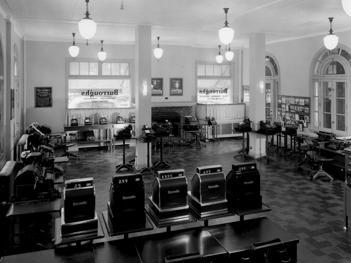 The image shows a vintage office with typewriters and other office equipment in a well-lit room with large windows and elegant light fixtures.