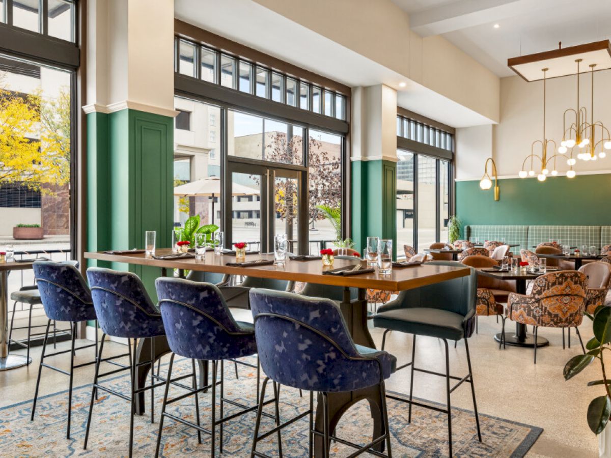 The image shows a modern restaurant interior with elegant lighting, green accents, and large windows, featuring tables and upholstered chairs.