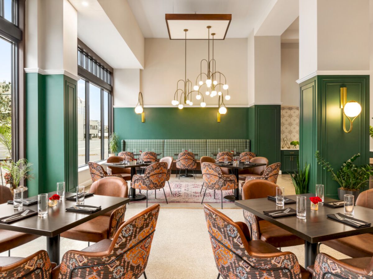 The image shows a modern restaurant with patterned chairs, green walls, and pendant lighting, creating a stylish and inviting atmosphere.