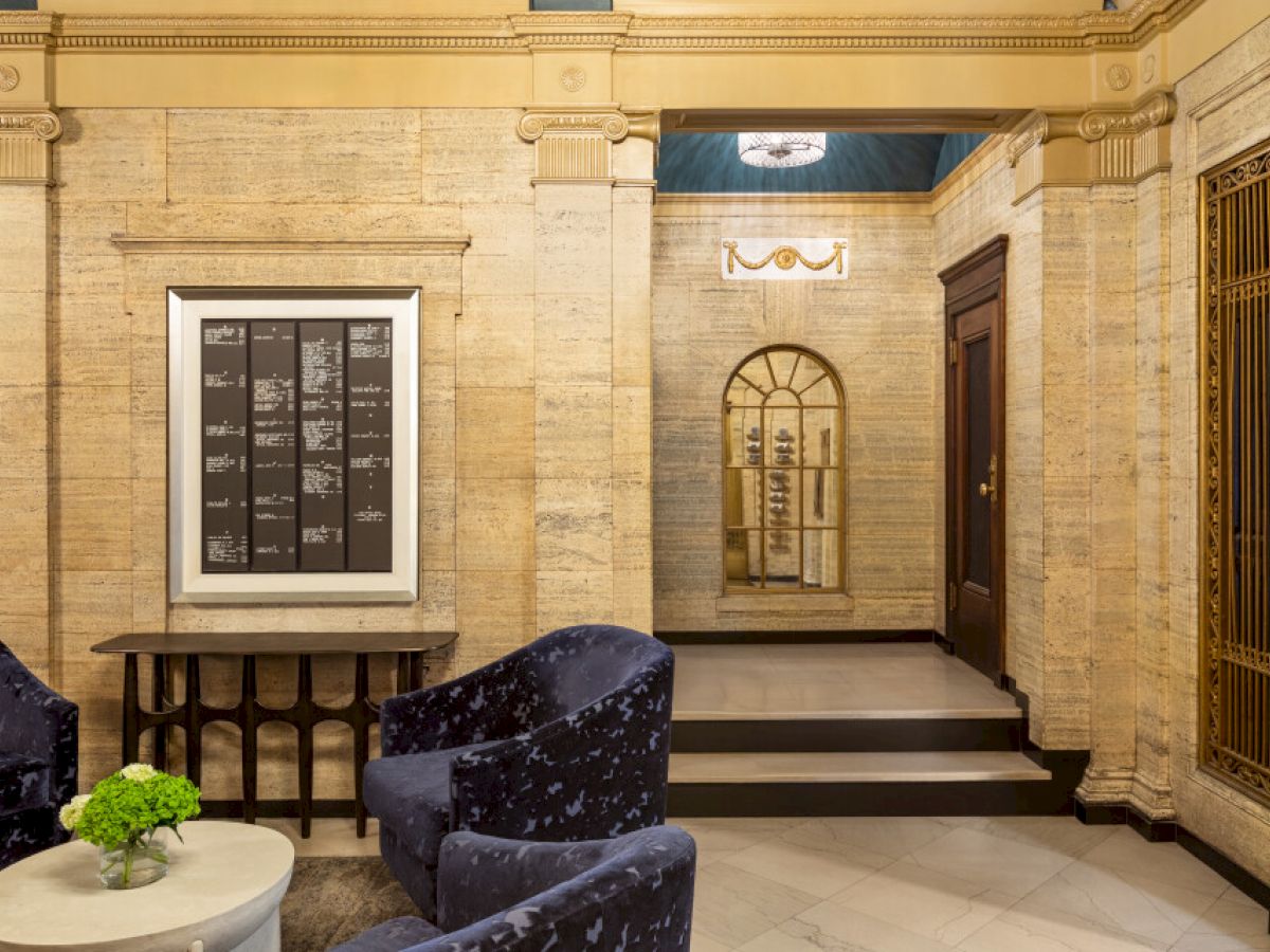 The image shows an elegant lobby with blue chairs, a side table with a plant, framed art on the wall, and a decorative mirror.