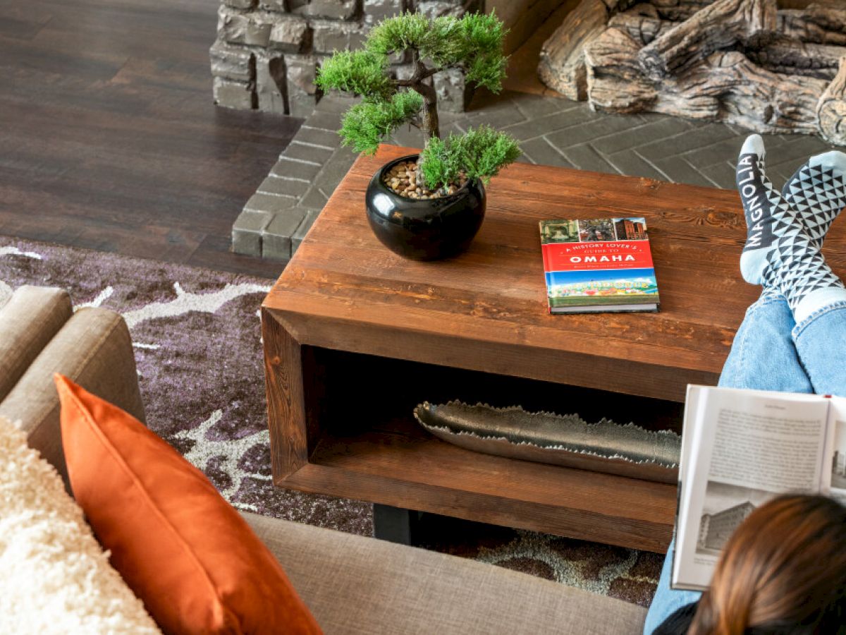 A cozy living room with a person reading, feet up on a table. A plant and a book are on the table near a stone fireplace and rug.