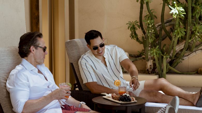 Two people are lounging on deck chairs outdoors, enjoying drinks with a tray of snacks nearby, next to a potted plant.