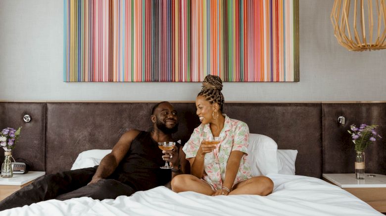 A couple is sitting on a bed, smiling and holding drinks. There's a colorful striped artwork on the wall behind them.