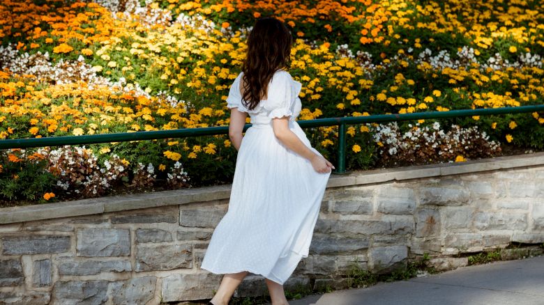 A woman in a white dress walks along a path beside a vibrant flower bed filled with yellow and orange blooms.