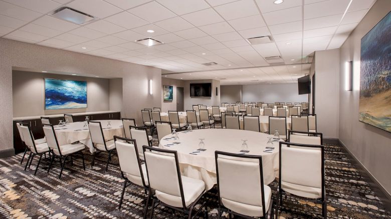 A conference room with round tables and white chairs, decorated with artwork and carpeted flooring, is arranged for a business event.