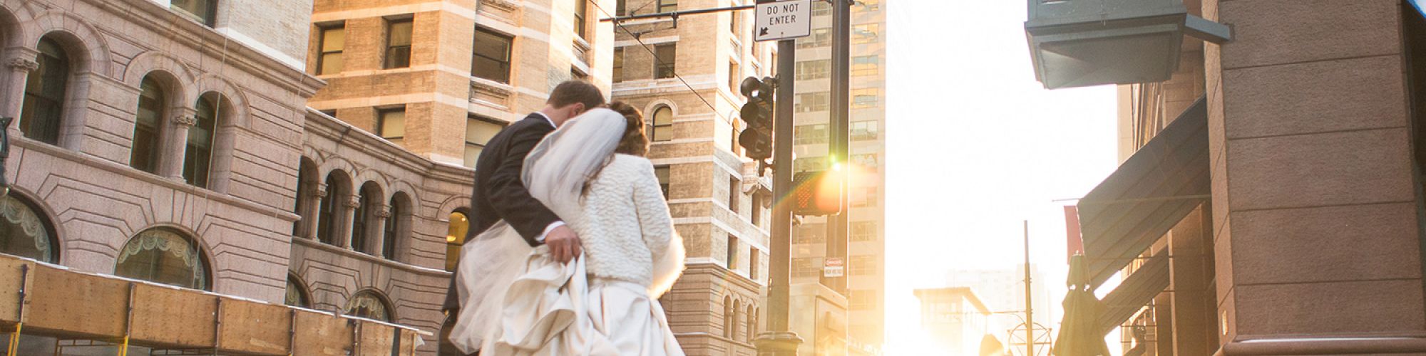 A couple in wedding attire embraces on a snowy street as the sun sets, beside a clock labeled 