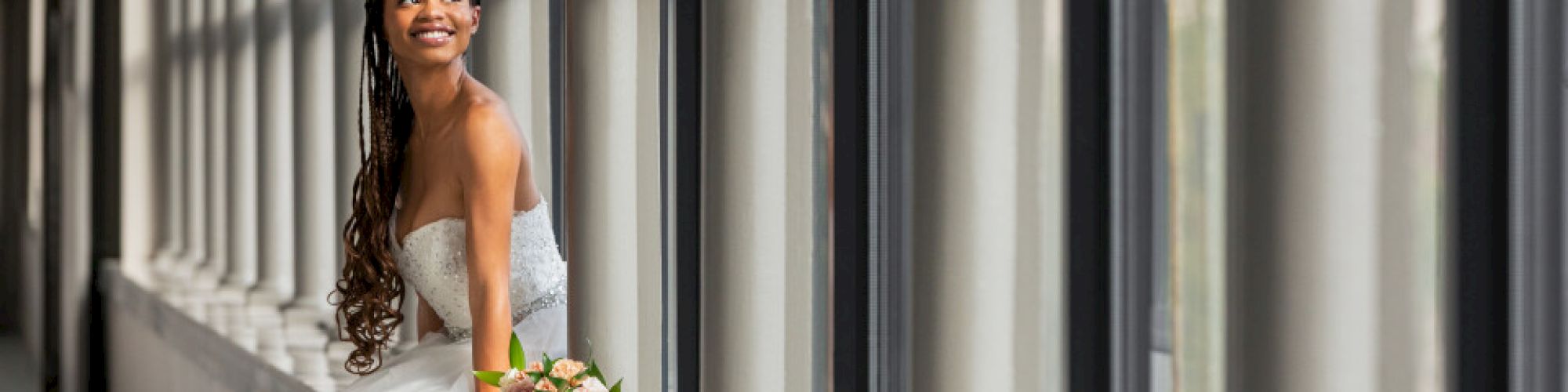 A woman in a wedding dress smiles while sitting by a row of windows, holding a bouquet of flowers.