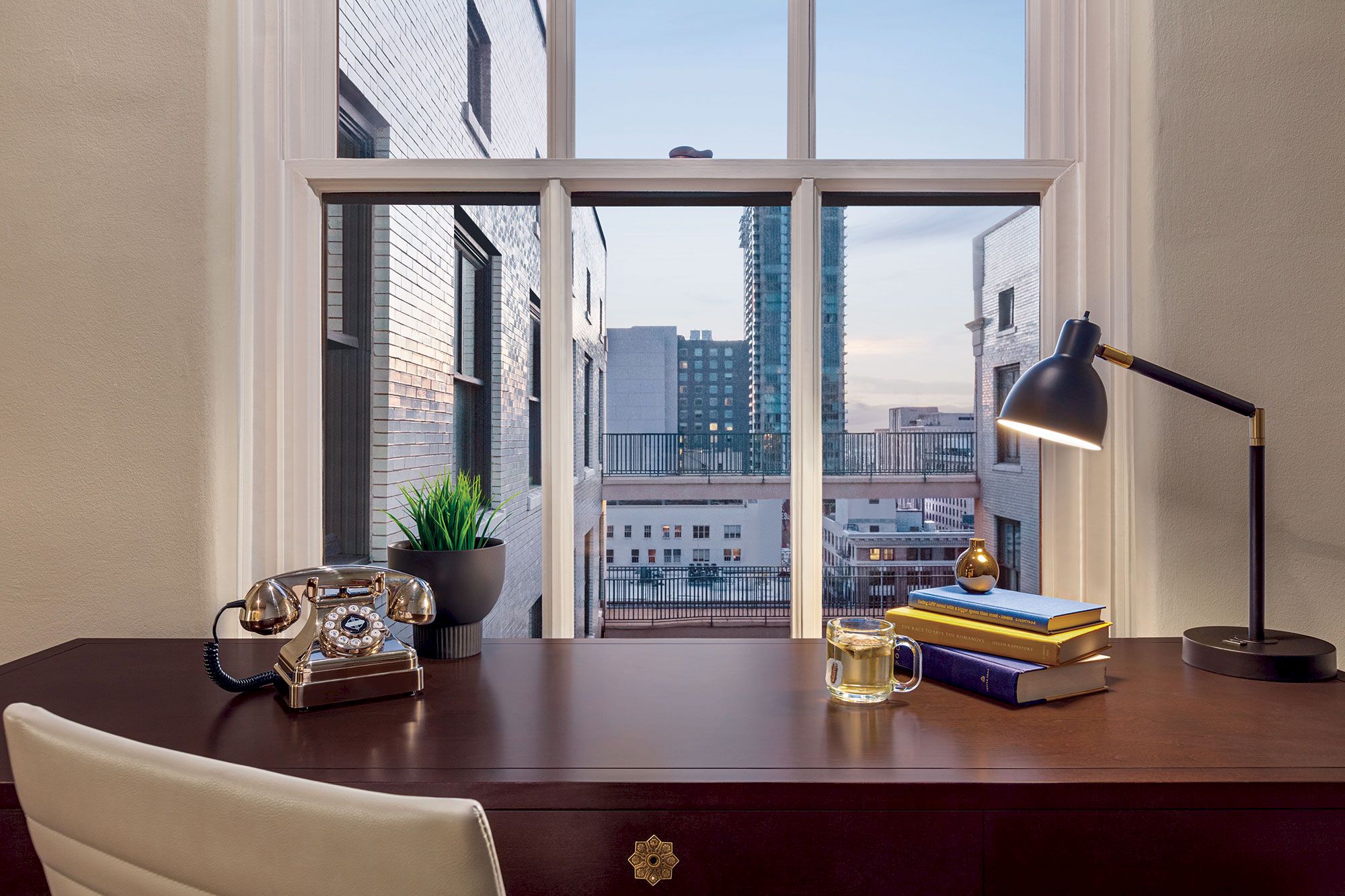 A desk with a lamp, books, a mug, and a vintage phone sits in front of a window overlooking city buildings and a clear sky.