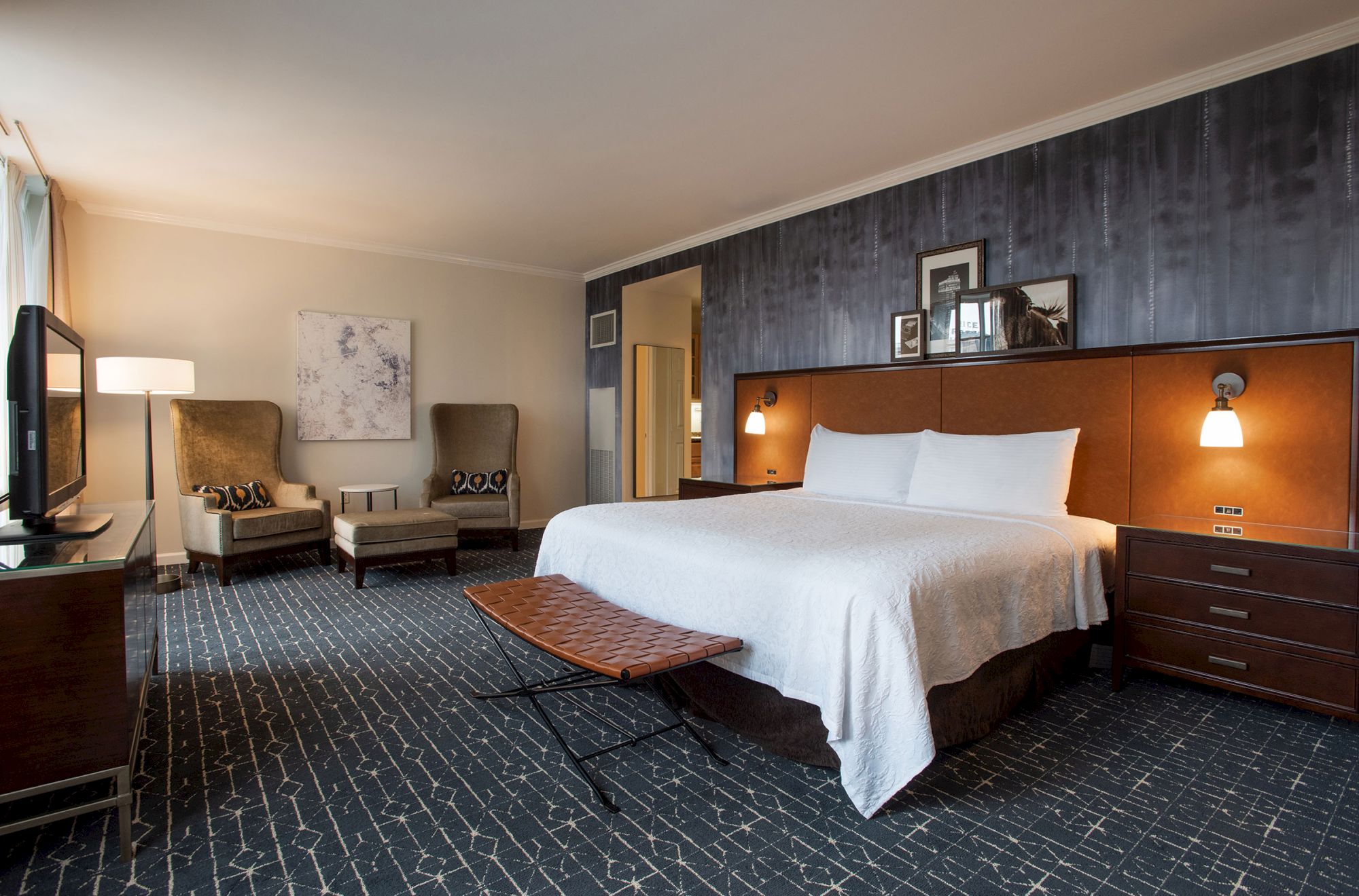 The image shows a modern hotel room with a large bed, two chairs, a TV, and stylish decor in neutral tones.