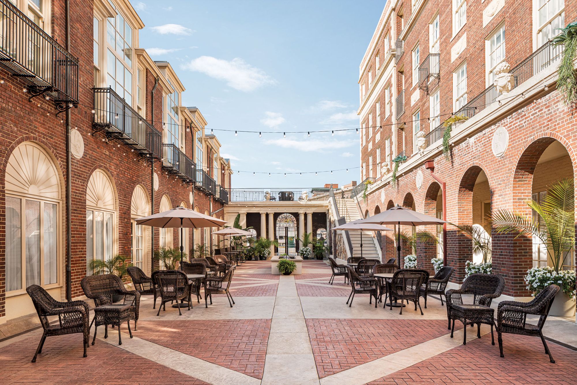 A charming brick courtyard with arched openings, outdoor seating, umbrellas, string lights, and potted plants under a clear blue sky.