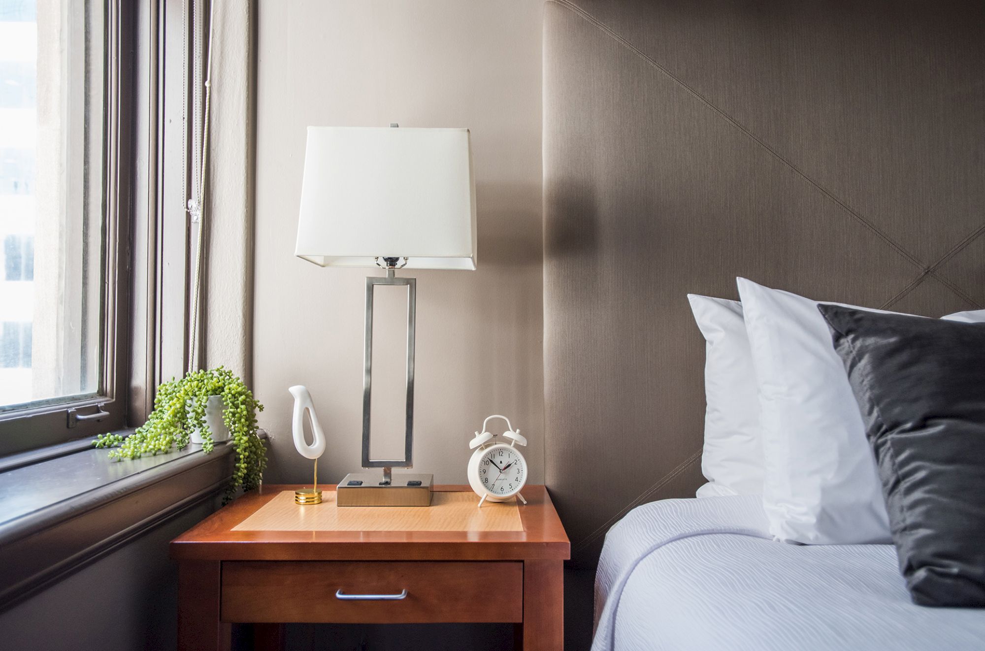 A cozy bedroom scene with a wooden nightstand, a table lamp, an alarm clock, a potted plant, and a neatly made bed with pillows.