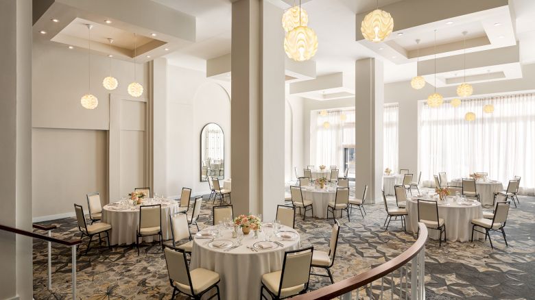 A spacious banquet hall with round tables, white tablecloths, floral centerpieces, and elegant pendant lights, ready for an event.