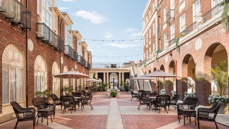 The image shows an elegant courtyard with brick buildings, outdoor seating, umbrellas, and string lights under a clear blue sky.