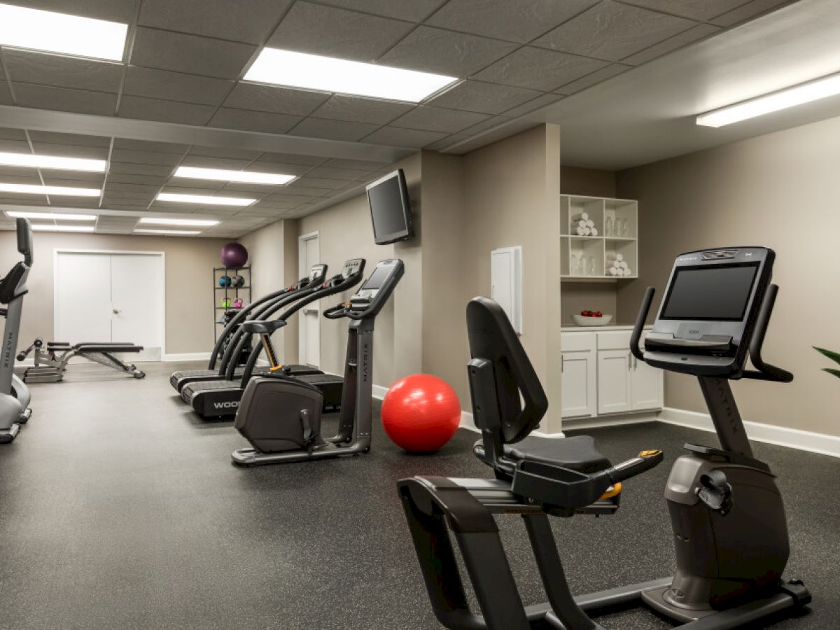 A gym room with exercise bikes, treadmills, a red exercise ball, a bench, and shelving, under bright ceiling lights.