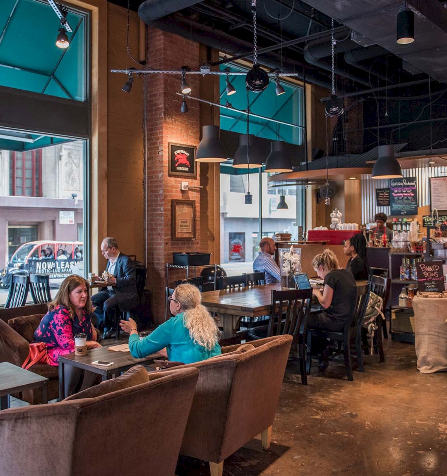 A cozy cafe with brown furniture, people talking and working, and a menu displayed above the counter. Large windows reveal street views.