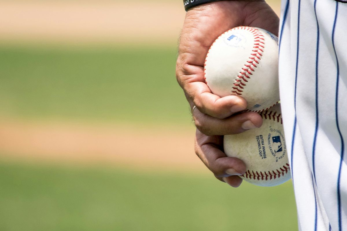 A person wearing a striped jersey holds two baseballs in their hand, with a grassy field in the background.