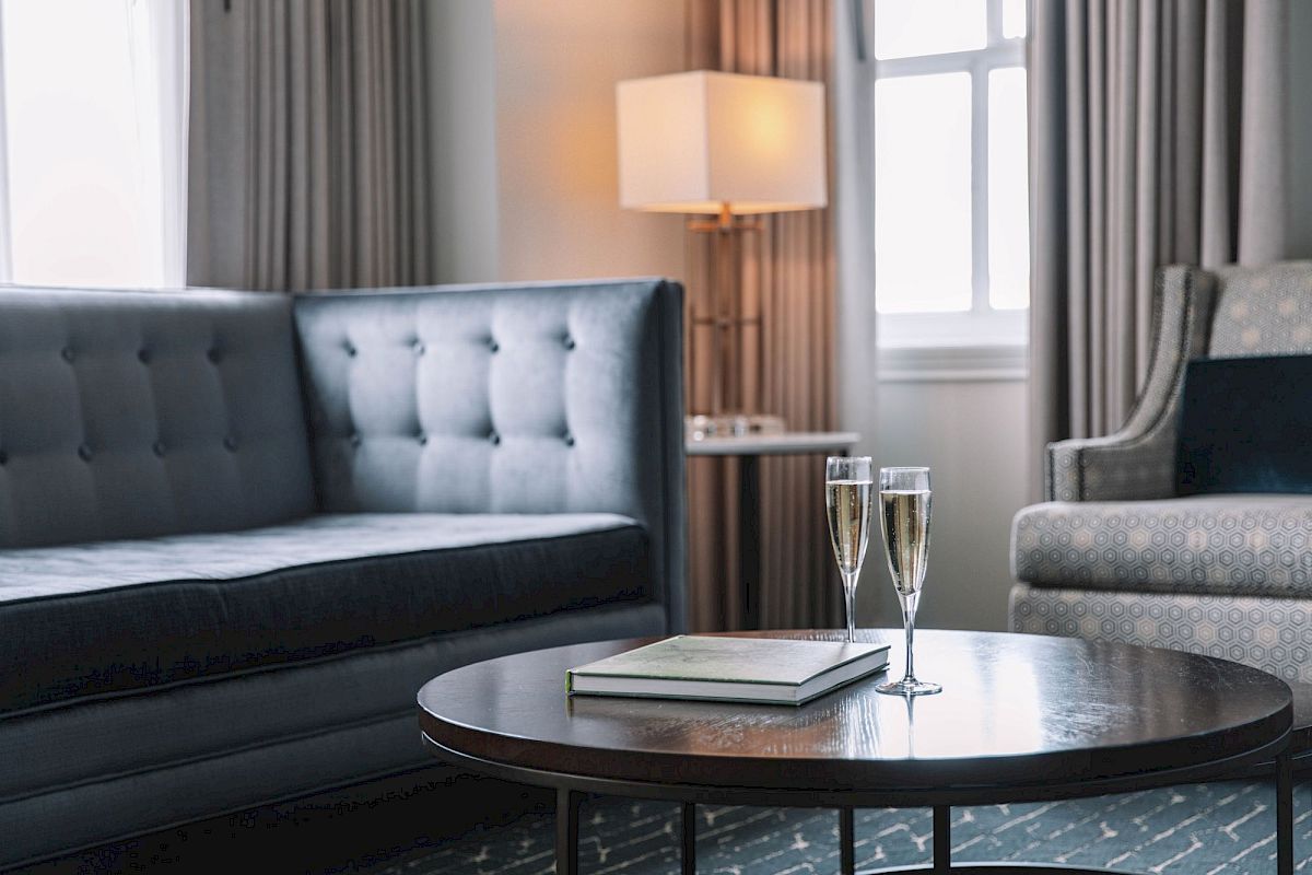 A cozy living room with a grey sofa, chairs, a round table with two champagne glasses, and a book, lit by a lamp near a window.