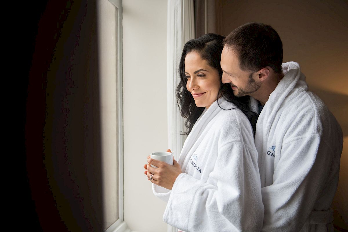 A couple in white robes is standing by a window; one holds a mug, both appear relaxed and content in a cozy setting.